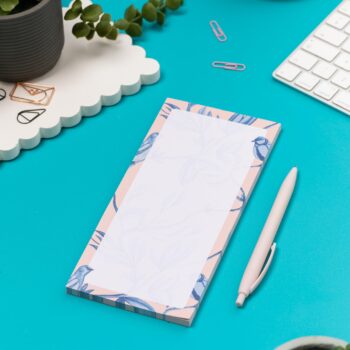 Brighton Birds List pad - Blush pink background with shades of blue bird and plant hand-painted motifs - is on a teal desk, next to a white pen, white keyboard and scattered paperclips.