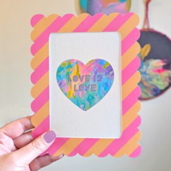 A close-up shot shows a hand holding a framed rainbow heart mini art print. The print features a colorful heart with "LOVE IS LOVE" text, central to the design. A pink and peach striped, scalloped frame surrounds the artwork. In the blurred background, hand-painted abstract circular wall art hangs on a white wall.