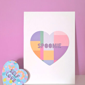 Pastel geometric heart art print with the word “Spoonie” on a white card, styled on a small shelf against a pink background.