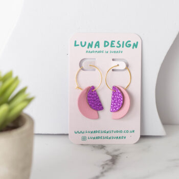 A pair of handmade earrings with gold hoops, featuring pink and glittery purple half-moon shapes, displayed on a "Luna Design" card. The earrings are set on a white marble surface next to a small potted plant.