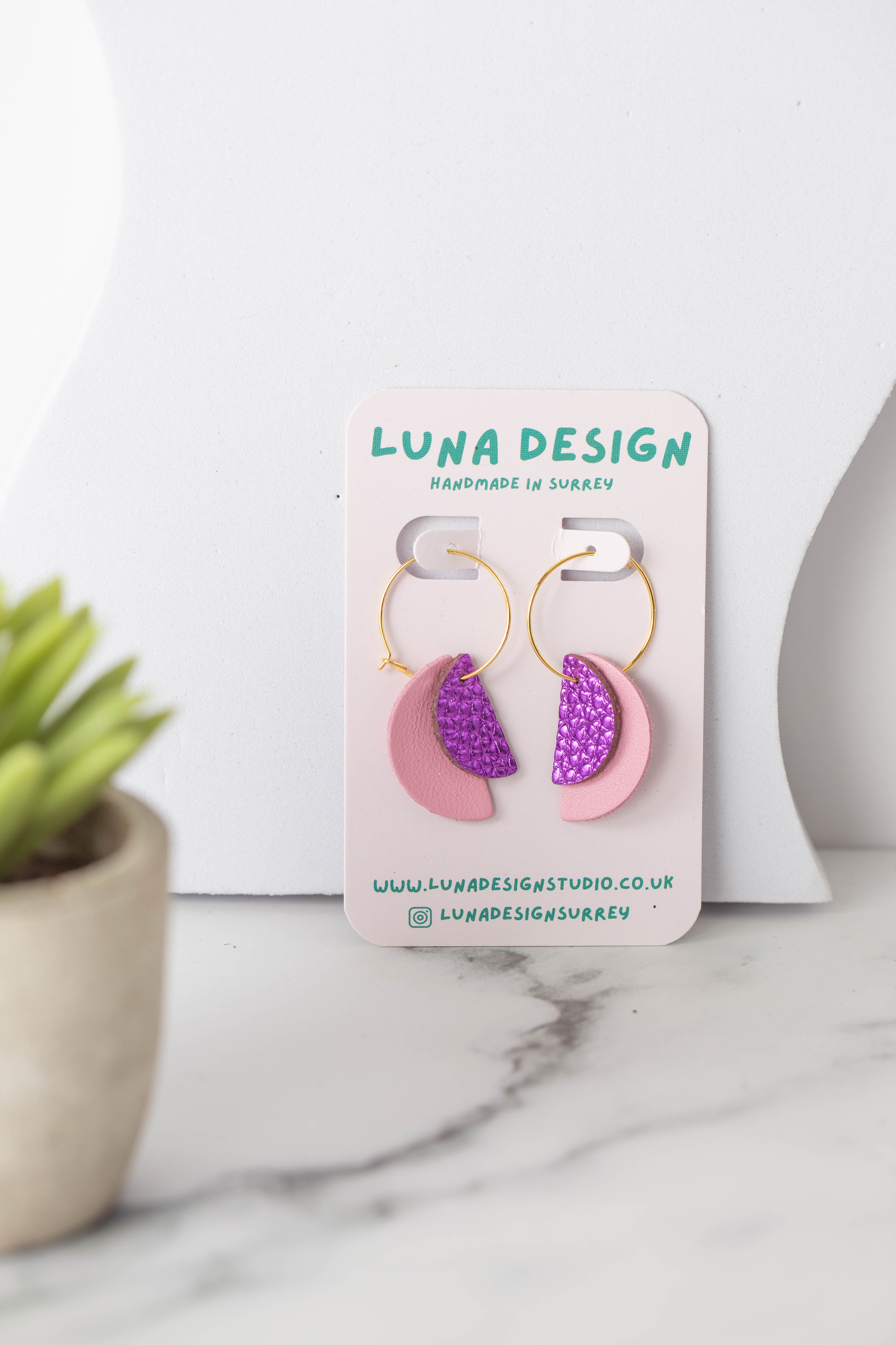 A pair of handmade earrings with gold hoops, featuring pink and glittery purple half-moon shapes, displayed on a "Luna Design" card. The earrings are set on a white marble surface next to a small potted plant.